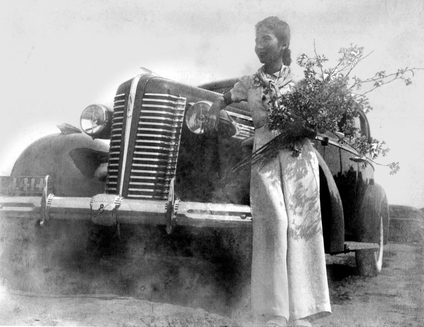 Madame Hoa Ly in ao dai holding an armful of blossom beside a 1938 Buick Century, in Da Lat