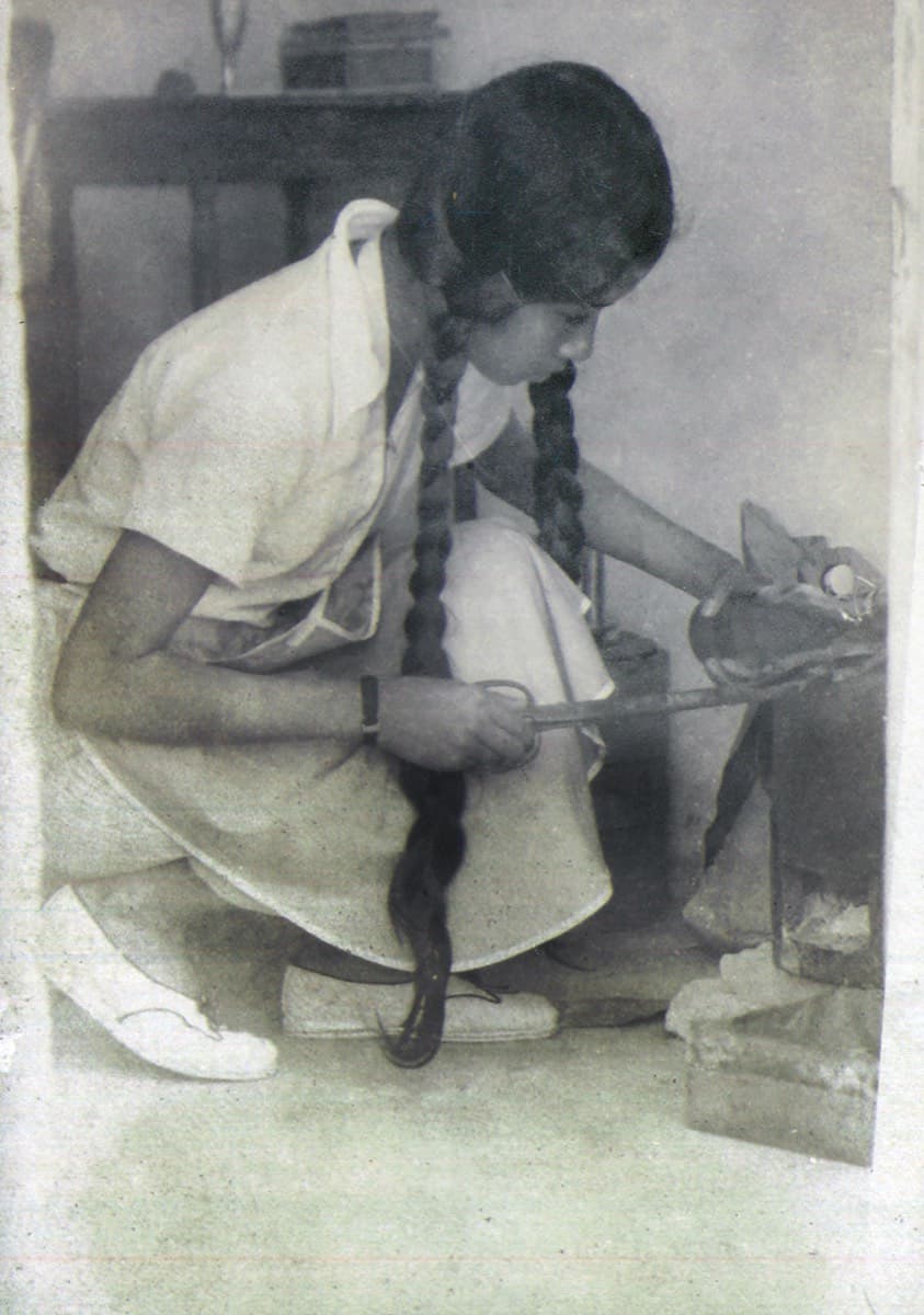 A girl in a white tunic with long plaits, crouched over a charcoal stove