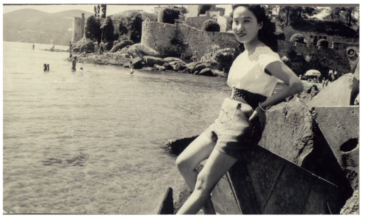Madame Hoa Ly leaning against a rock at the water's edge, a stone fort behind