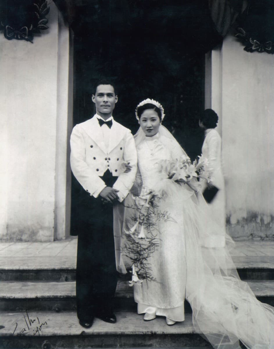 A bride in a white veil and long train beside a groom in a white dinner jacket, on stone steps