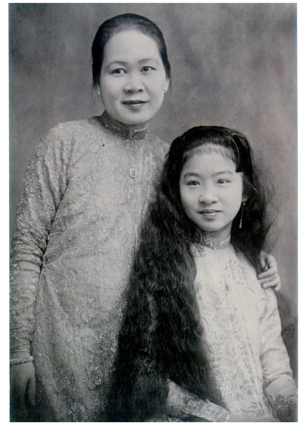 Madame Hoa Ly as a young woman, standing beside her seated mother, both in embroidered ao dai